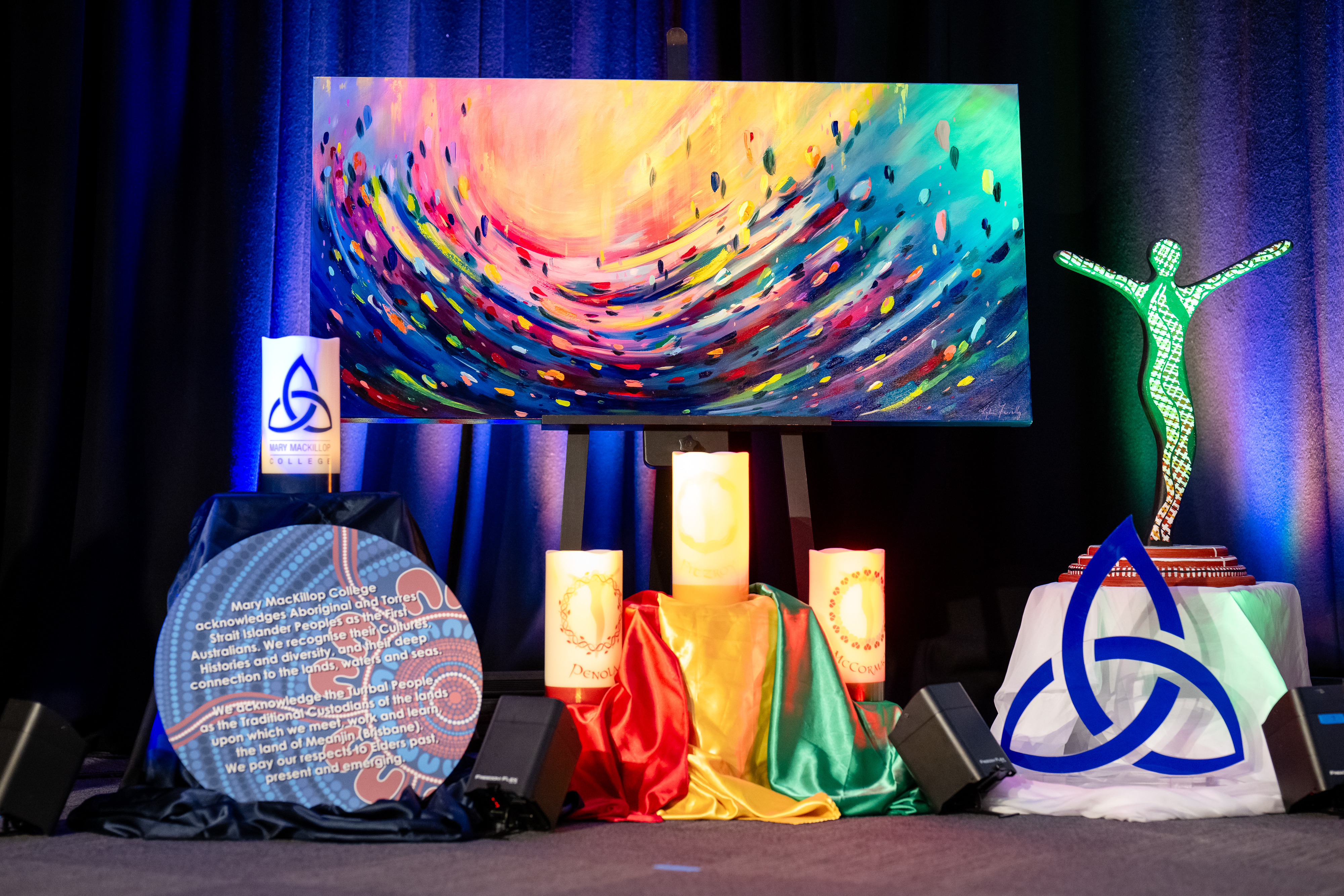 Display of religious artwork featuring a colourful abstract painting, lit candles, fabric draping and symbolic sculptures on a stage backdrop