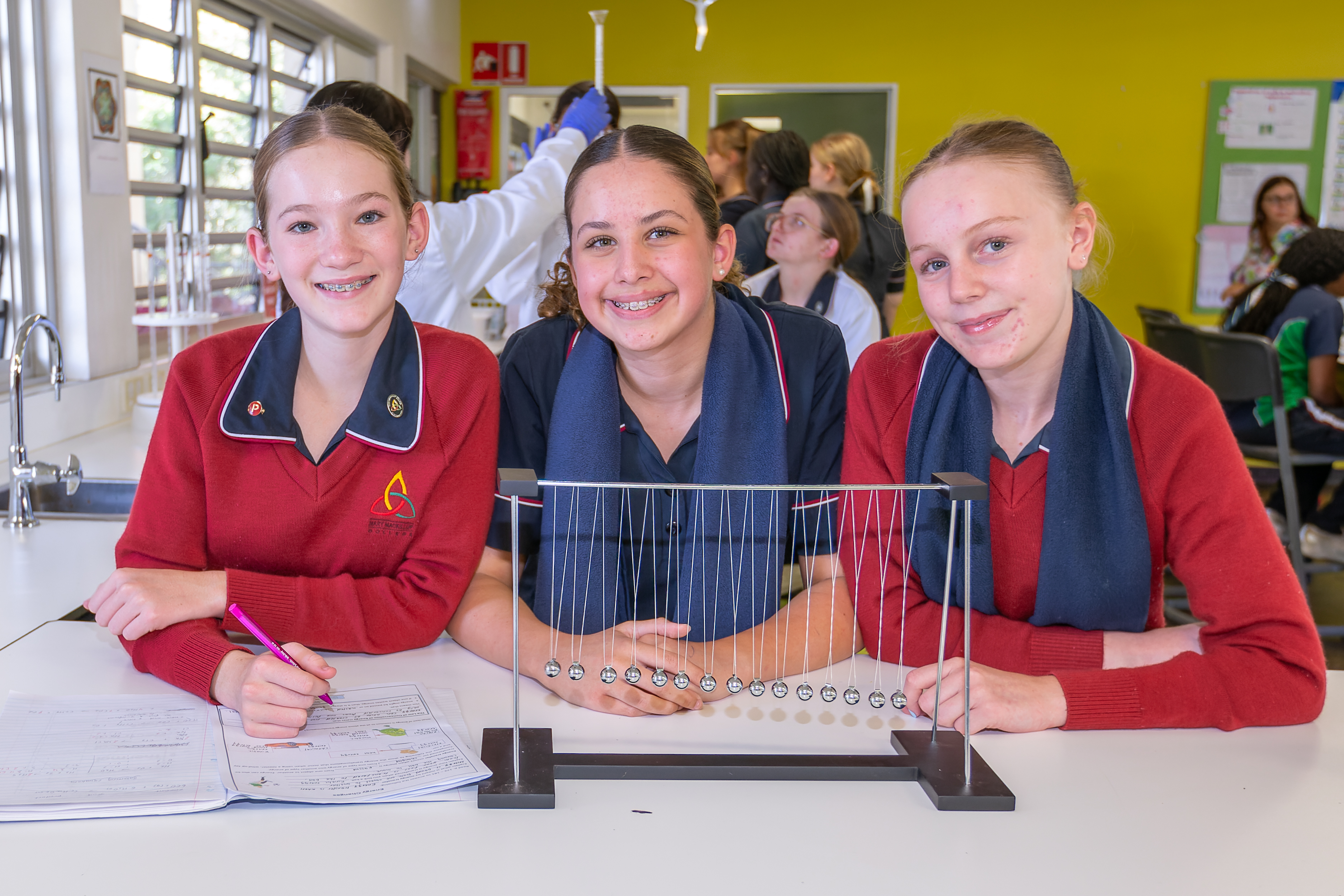 Three students in school uniforms sitting at a science bench with a Newton’s cradle and worksheets