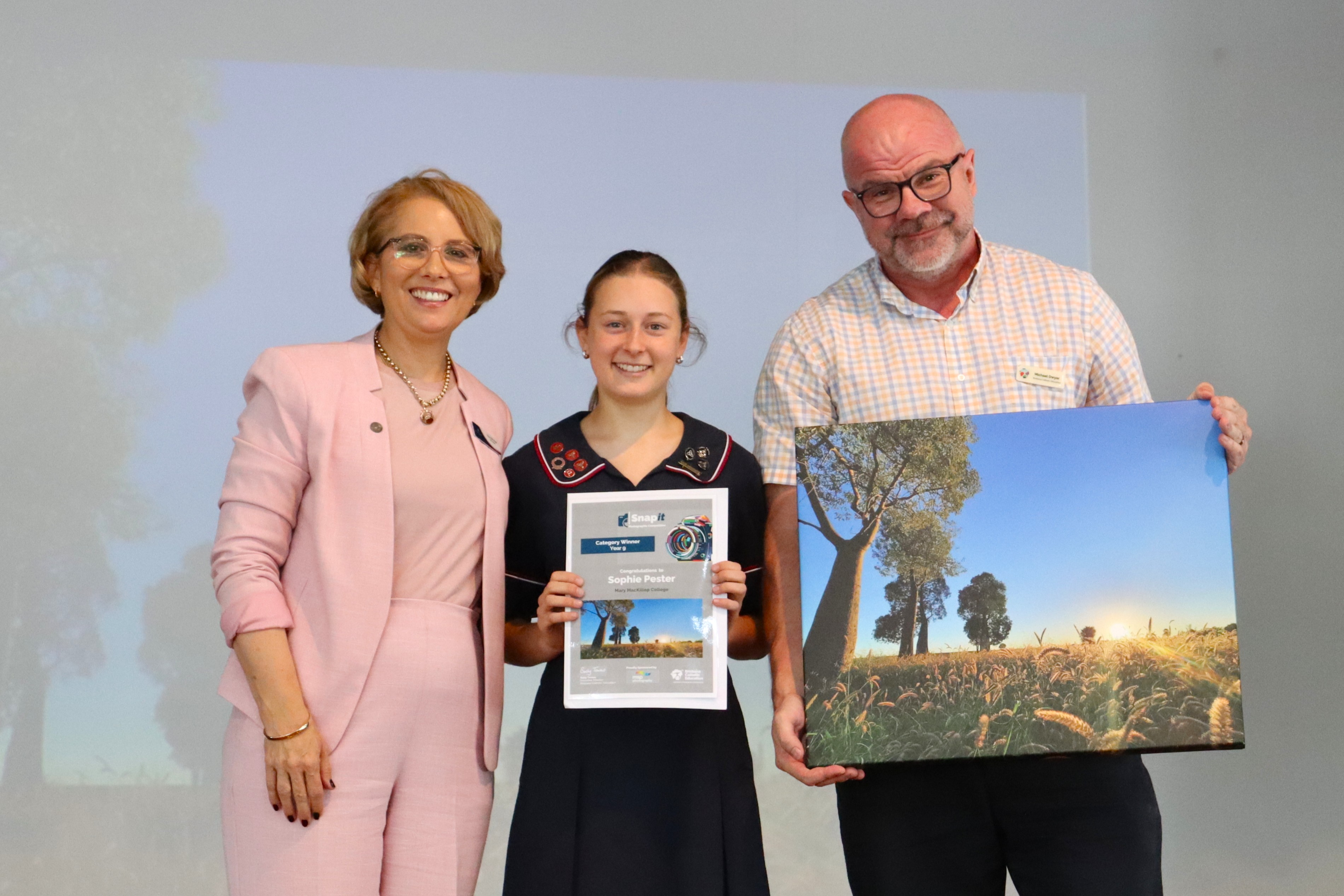 Student holding a certificate standing between two adults presenting an award and framed photograph