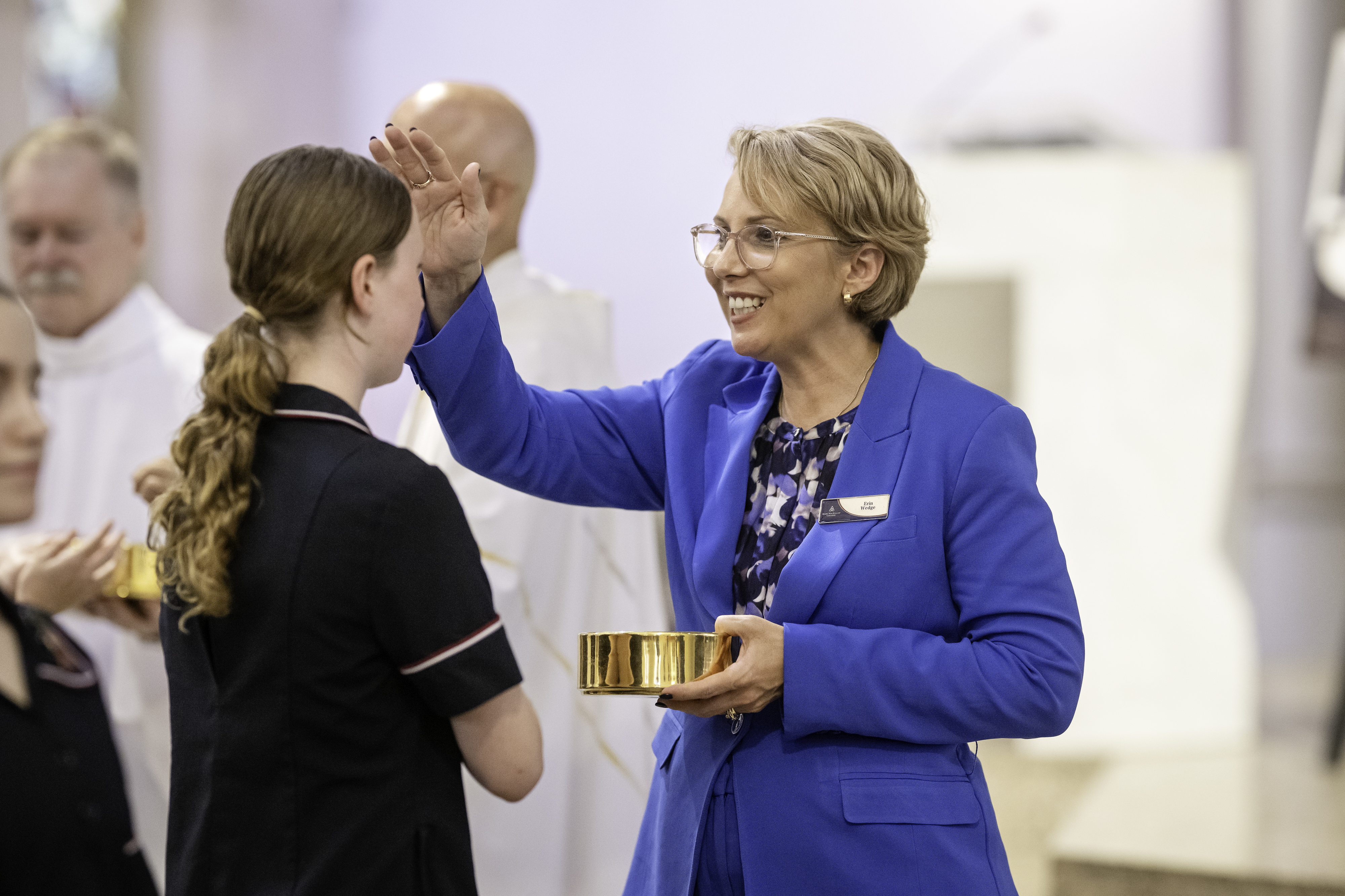 The Principal Adult marking a student’s forehead during a school liturgy while holding a small metal bowl
