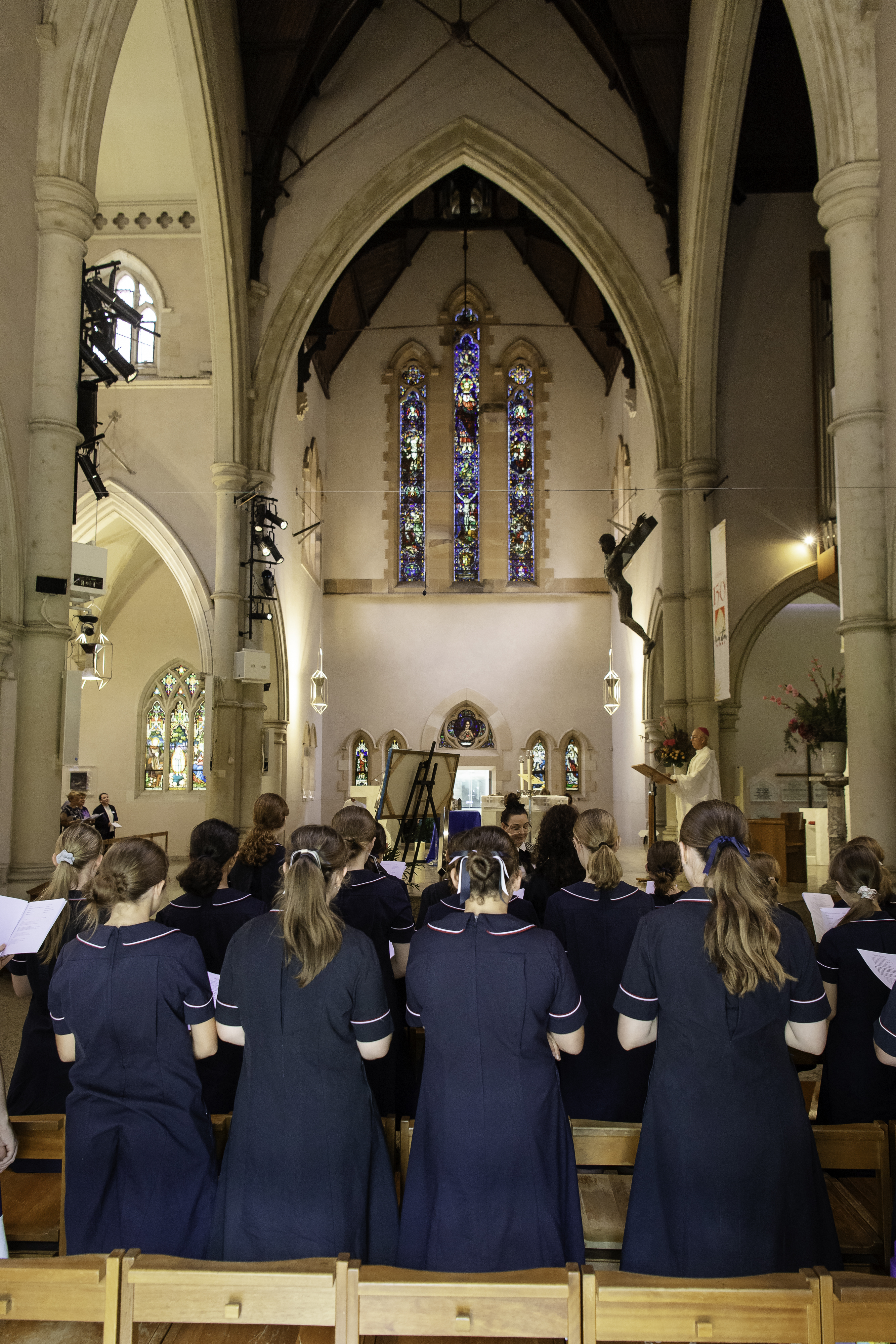 Students in school uniforms standing inside a church facing the altar with stained glass windows and religious artwork visible