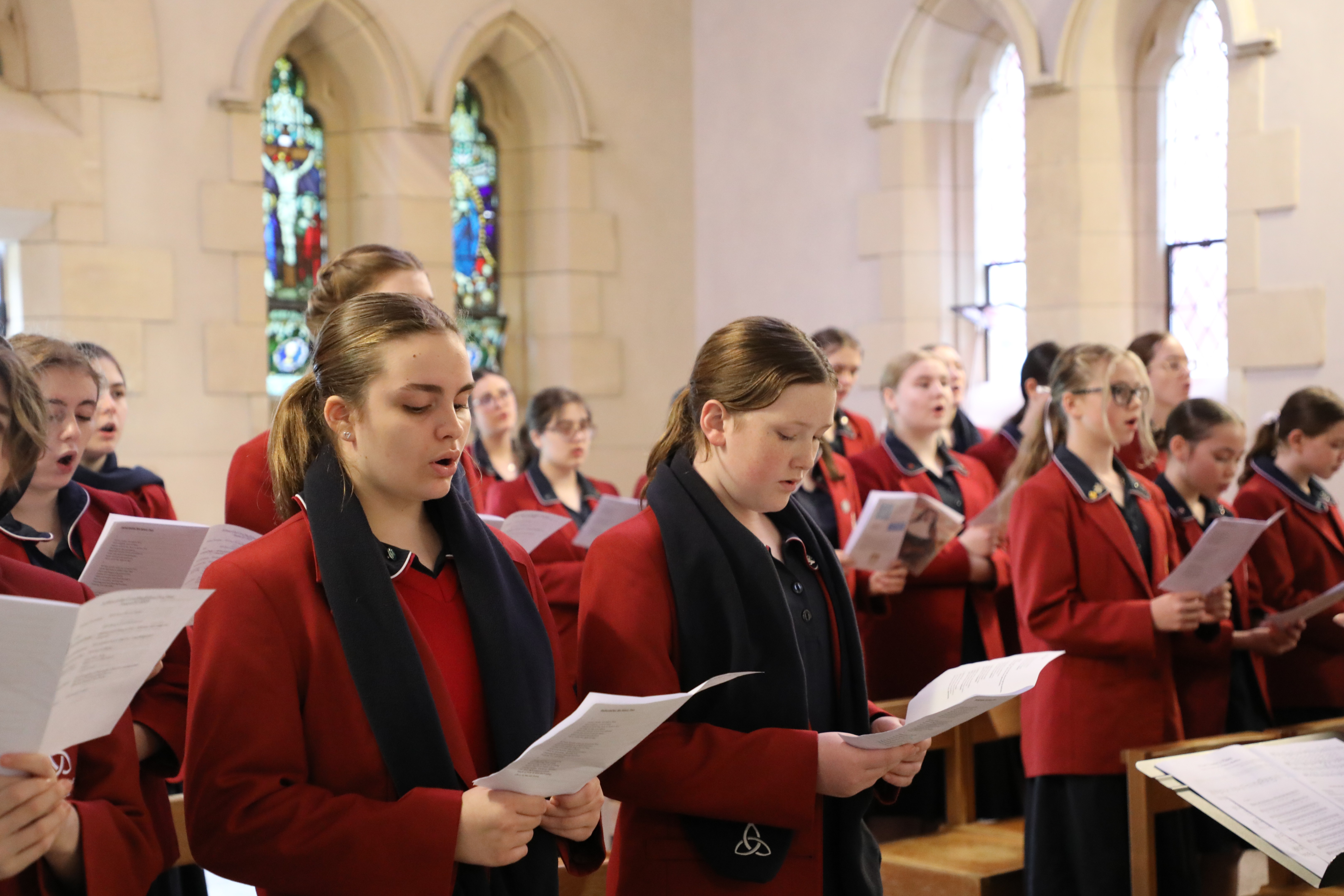 Students in school uniforms standing inside a church singing from printed sheets during a choir performance