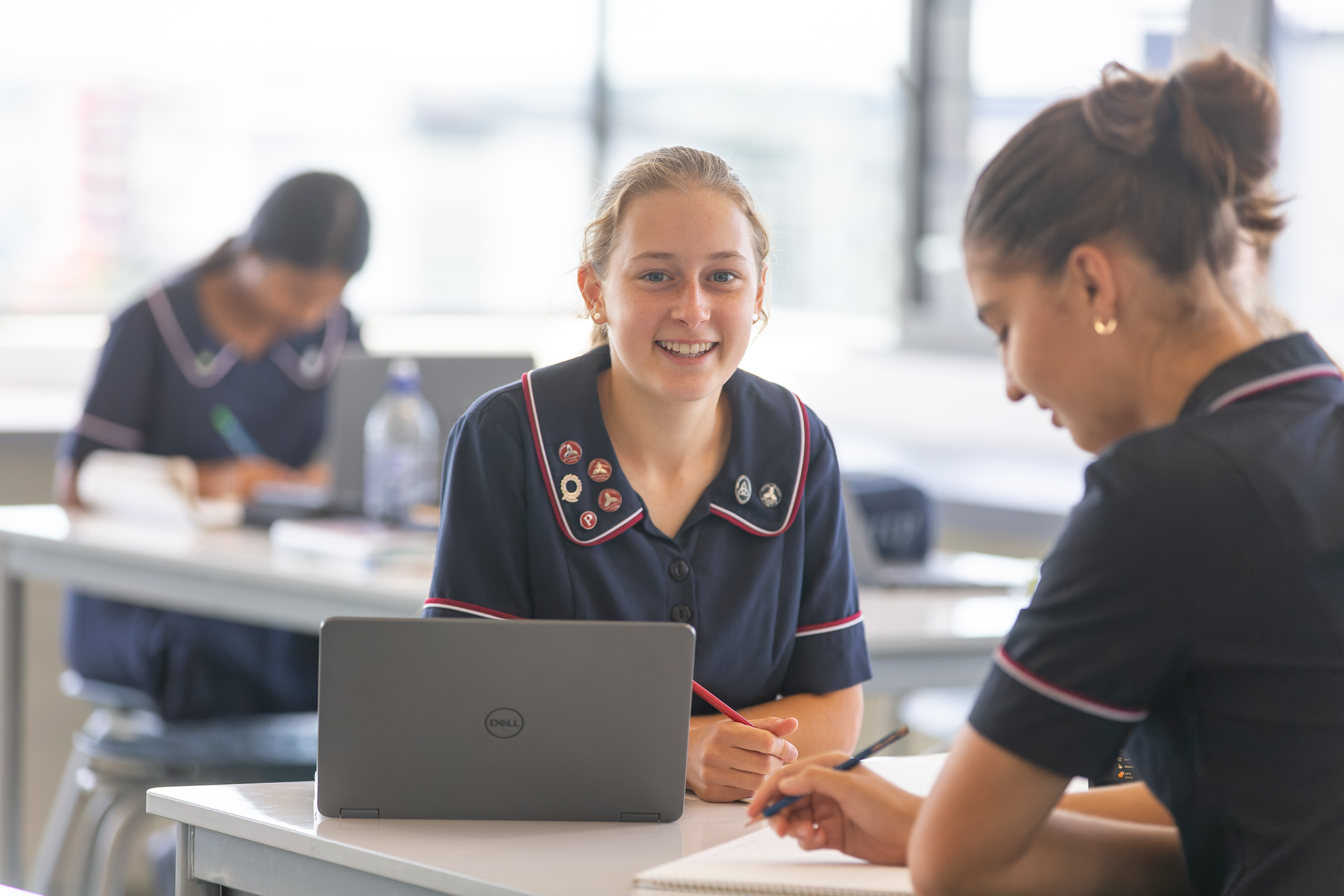 Student in school uniform sitting at a desk using a laptop while another student writes beside them in a classroom