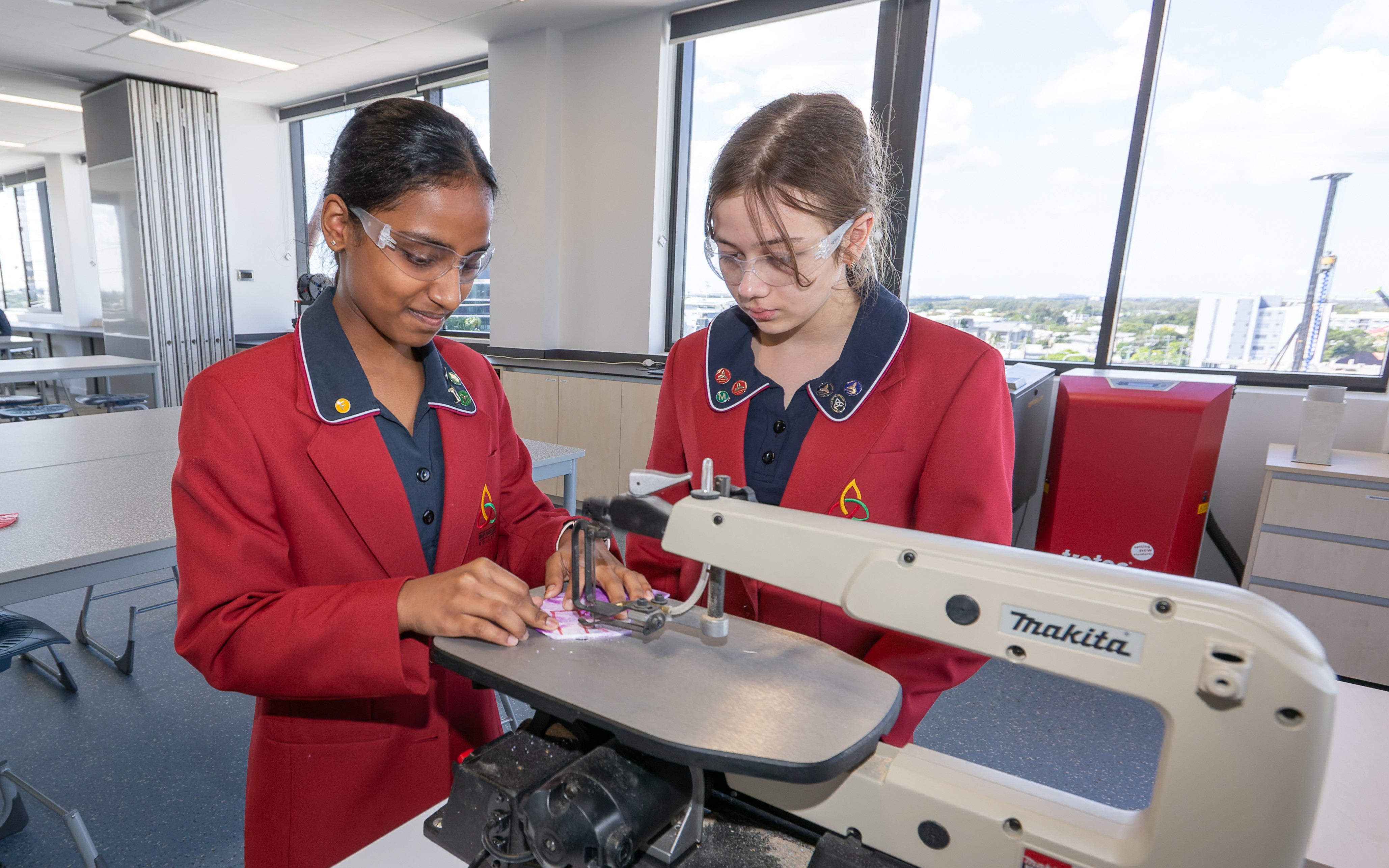 Two students in school uniforms wearing safety goggles working together on a scroll saw machine in a classroom workshop