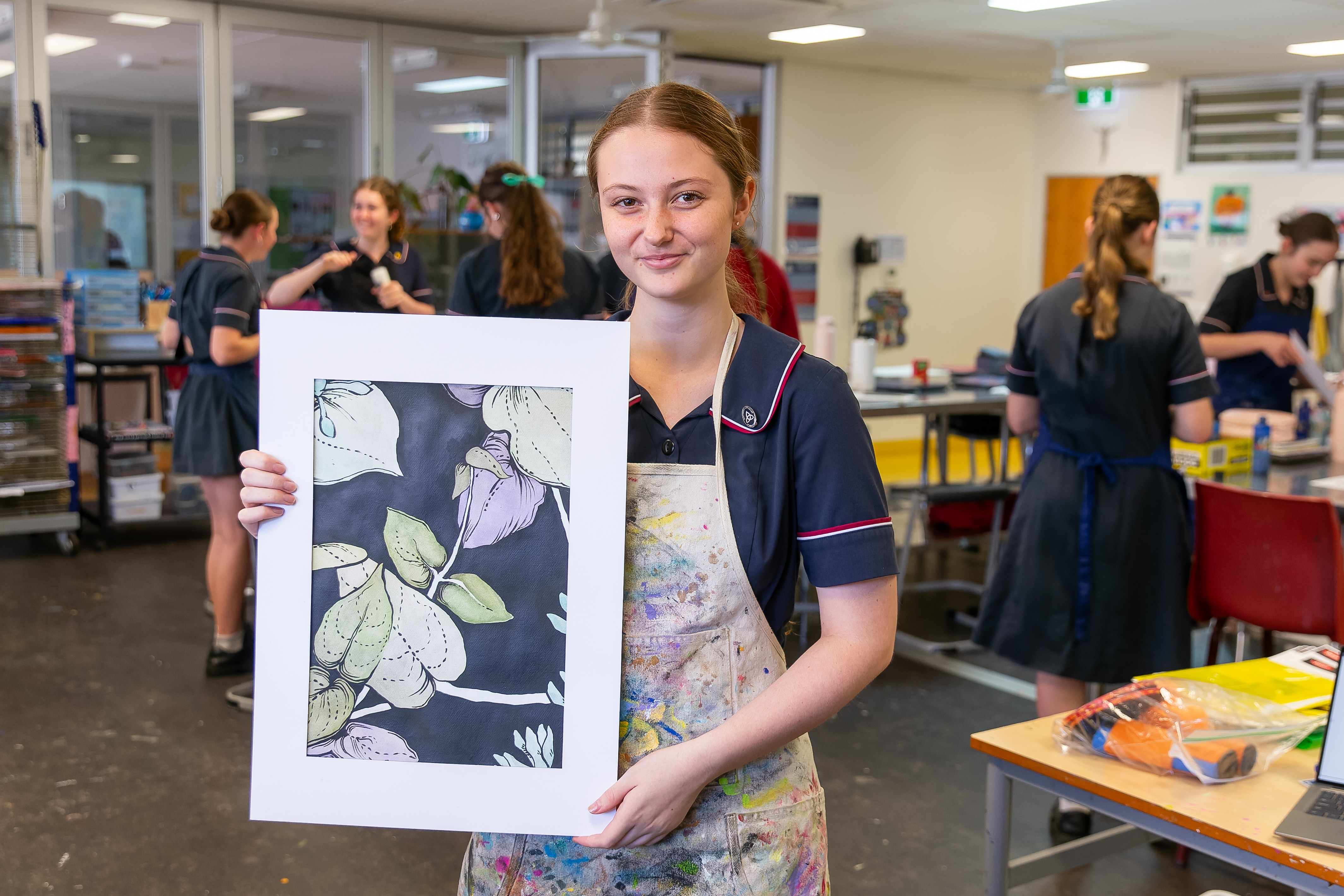 Student in school uniform holding a printed artwork while standing in an art classroom with other students working in the background