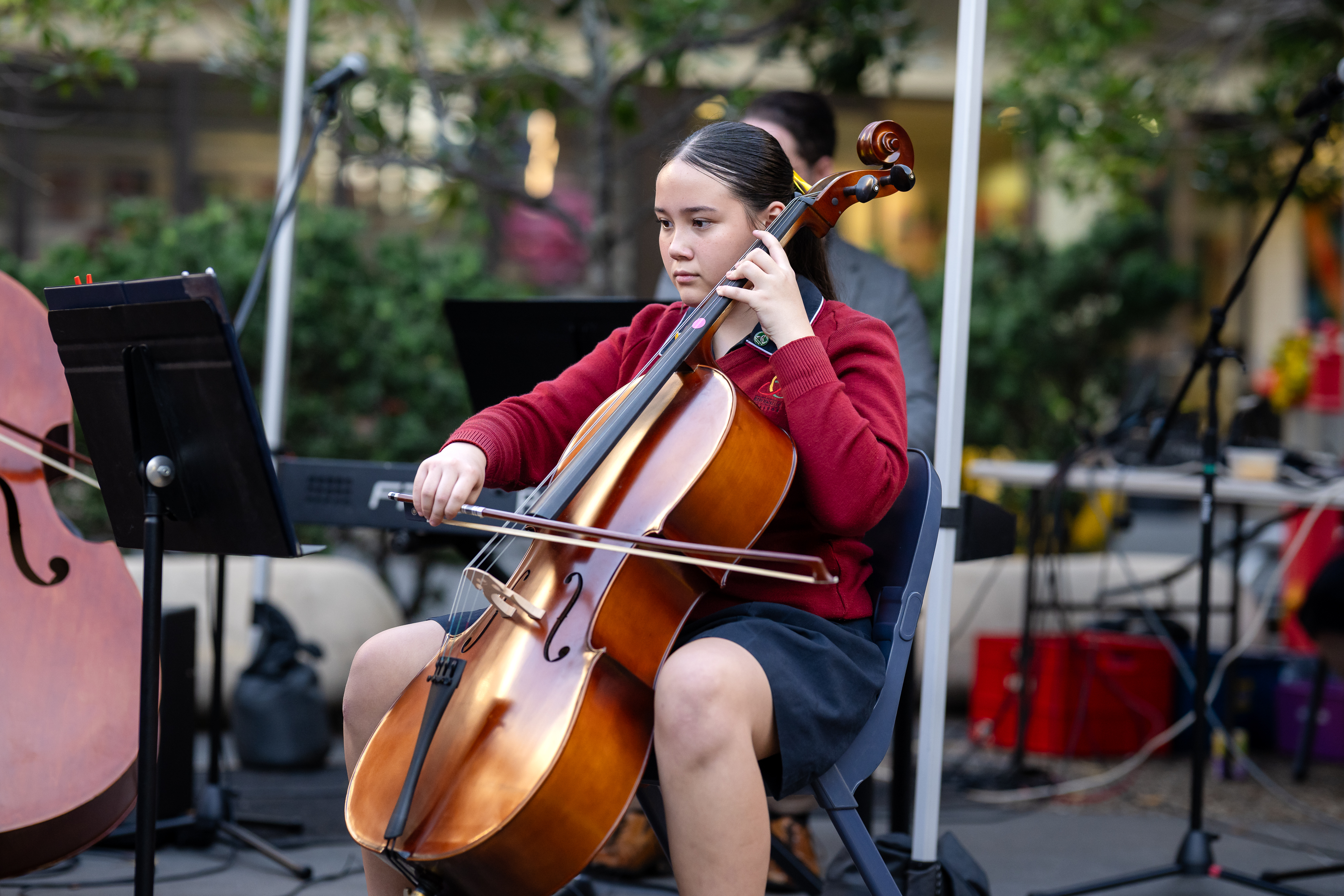 Student in school uniform seated and playing a cello during an outdoor performance