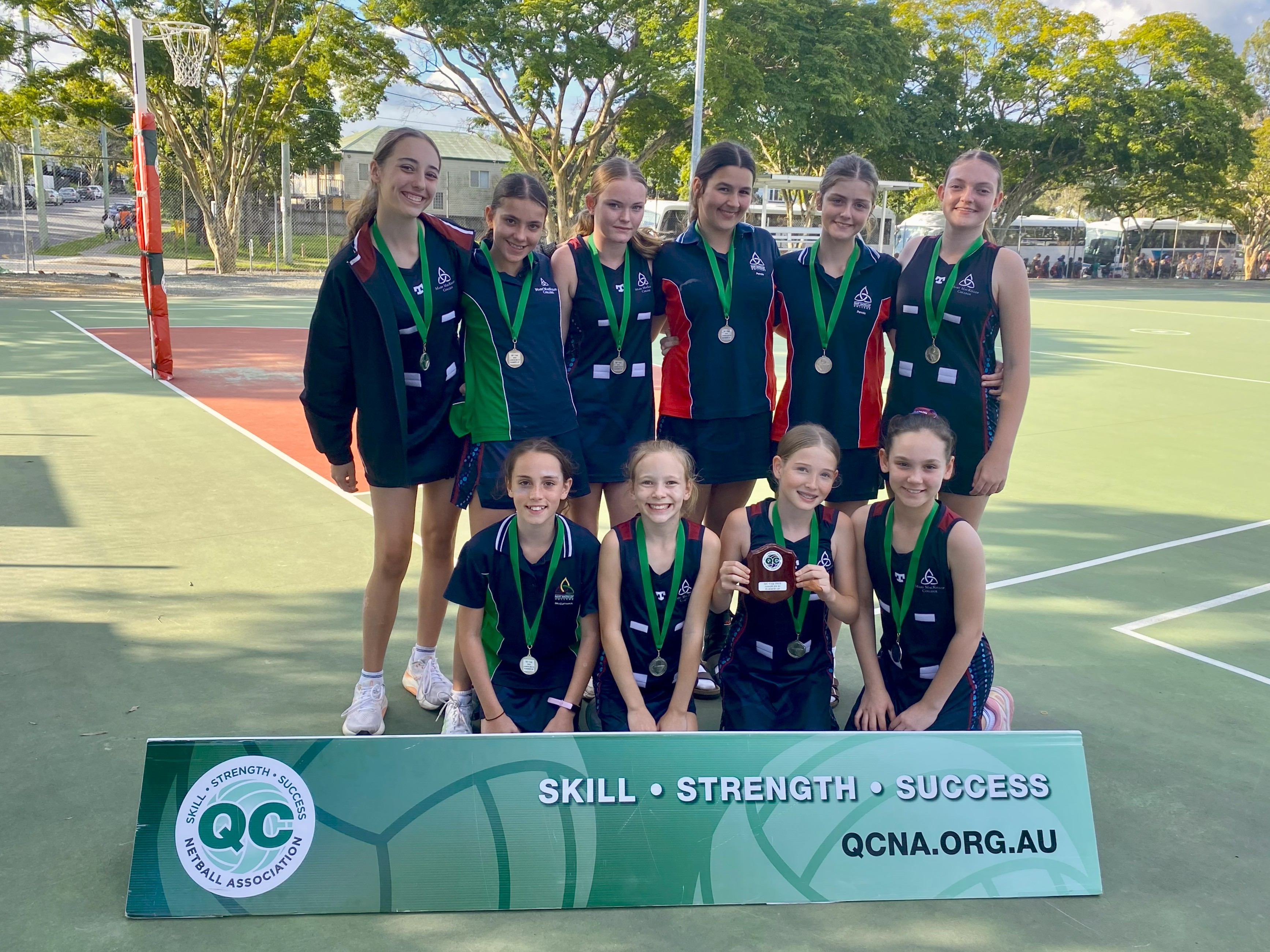 Students in school sports uniforms standing and kneeling on a netball court holding medals and a team trophy behind a QCNA competition banner