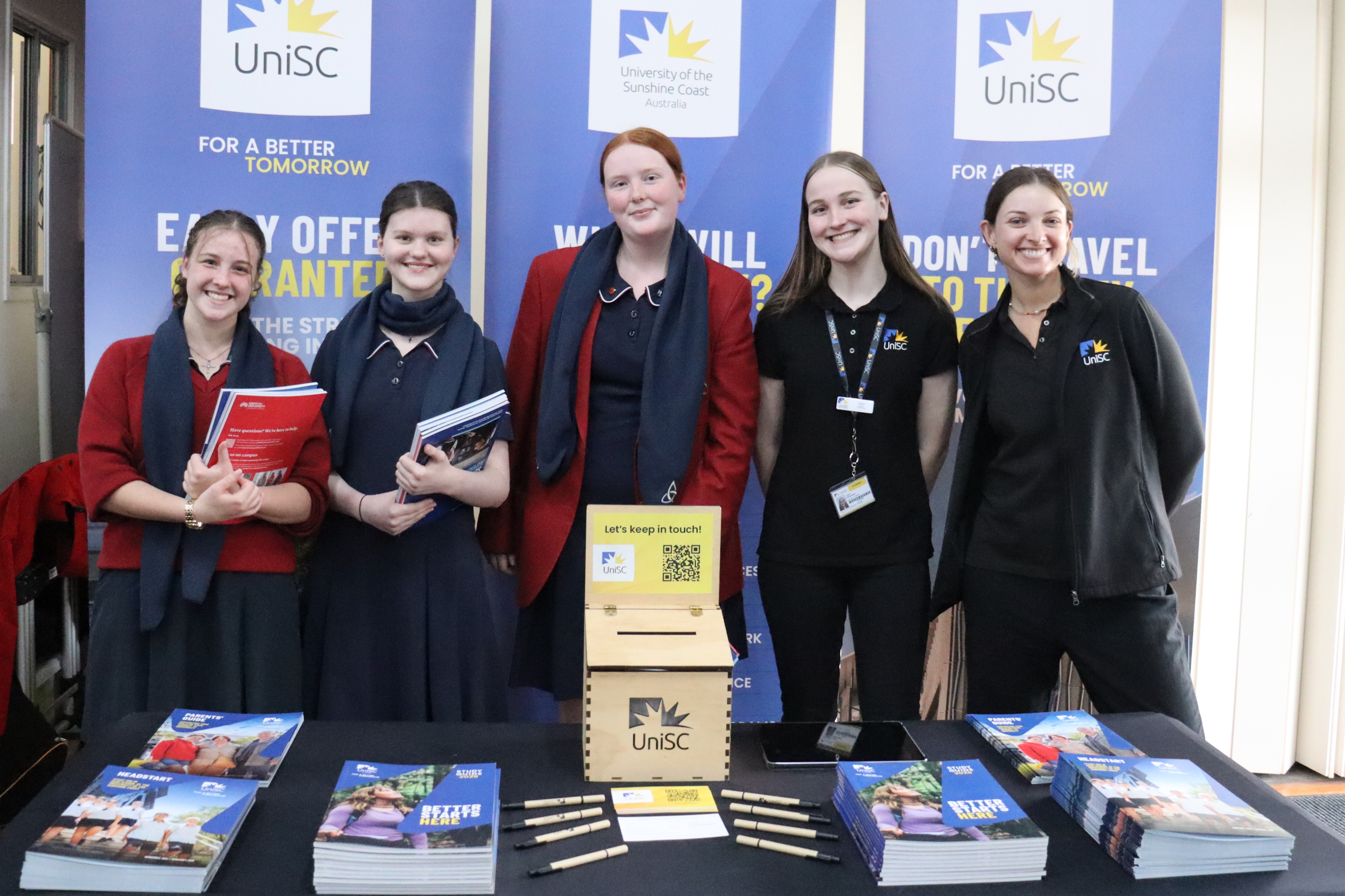 Group of students and staff standing behind a UniSC information table with brochures and banners