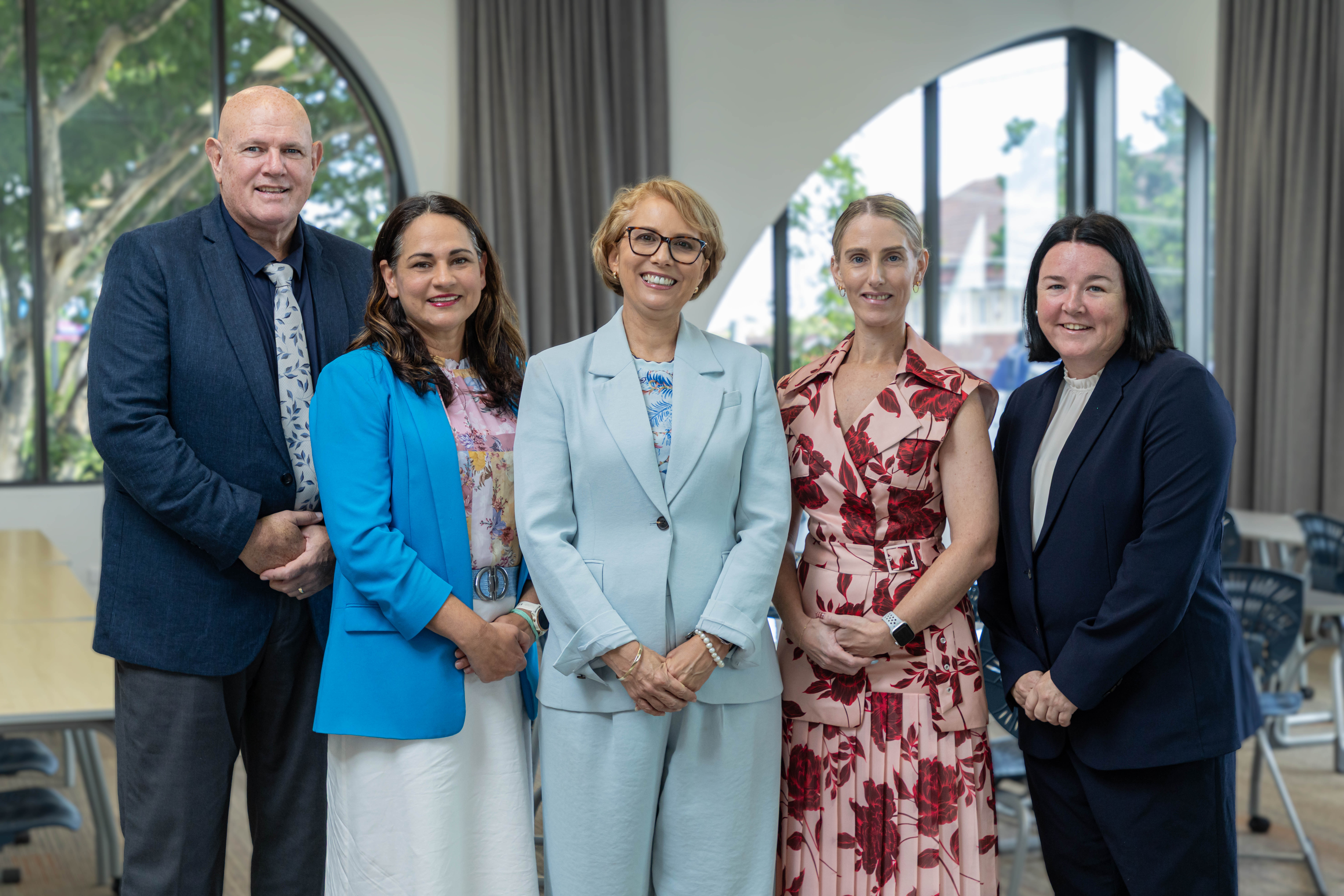 Group of five adults standing together indoors in a meeting room with tables and chairs behind